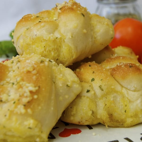 Golden-brown garlic knots, sprinkled with herbs and cheese, with tomatoes and spices in the background.