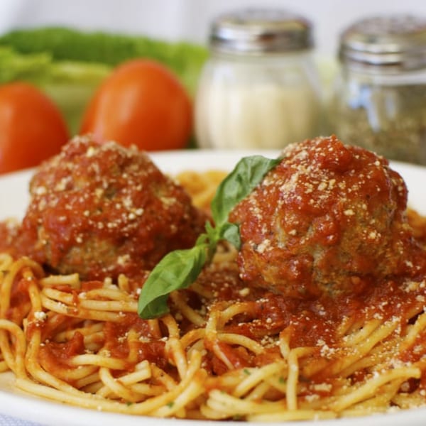 Spaghetti and meatballs with tomato sauce, parmesan, and basil, with tomatoes and shakers in background.